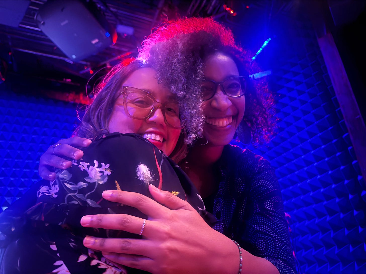 a light skinned miffle aged Puerto Rican woman with glasses embraces a black woman with glasses. Both smile joyfully into the camera