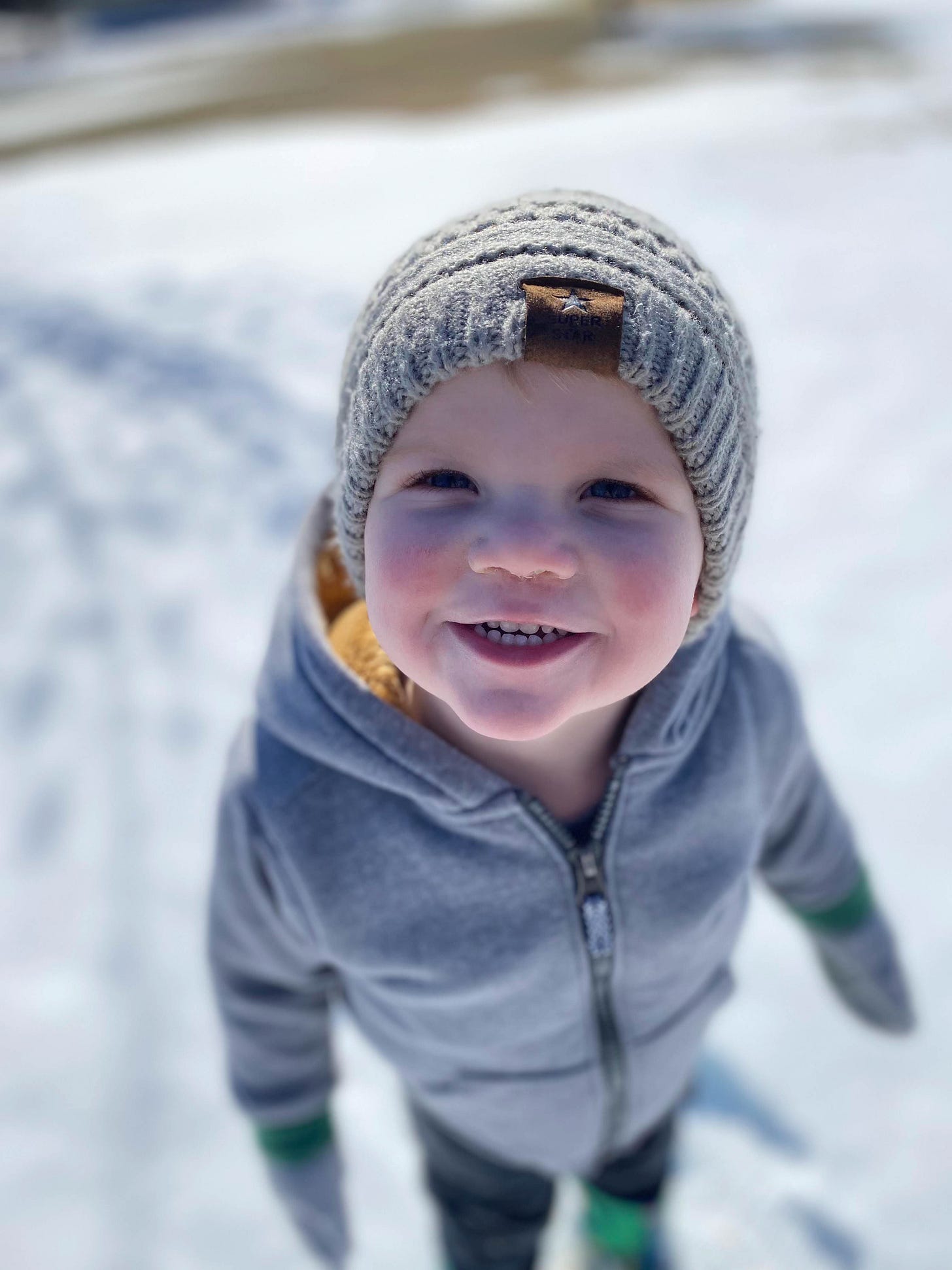 Author's grandson dressed in coat, hat, and mittens, smiling, with snow in the background.