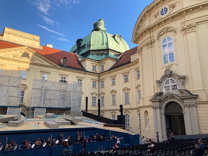 Title: UV-SOM-FD-01-Oper-Klosterneuburg-Stage-Imperial-Courtyard.jpg - Description: Open-air opera stage and orchestra inside the Baroque courtyard of Klosterneuburg Abbey, surrounded by historic architecture beneath a blue evening sky.