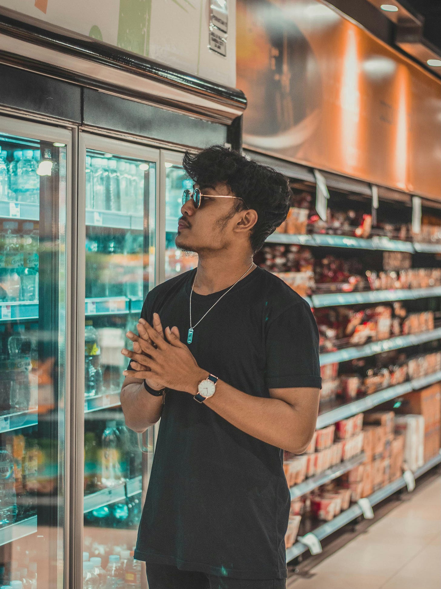 A young man with brown skin stands in a grocery store aisle, looking at the items inside a glass-doored refrigerator. He is wearing a black t-shirt, sunglasses, and a watch, with their hands clasped together in front of them. The aisle is well-lit, and shelves stocked with various products are visible in the background. A young man with brown skin stands in a grocery store aisle, looking at the items inside a glass-doored refrigerator. He is wearing a black t-shirt, sunglasses, and a watch, with their hands clasped together in front of them. The aisle is well-lit, and shelves stocked with various products are visible in the background.