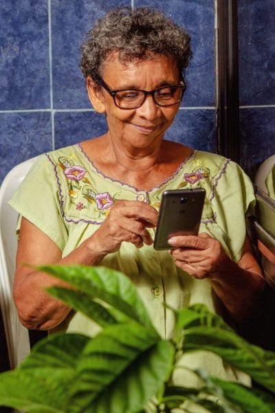 20260711_idosa_celular Uma idosa sentada em uma cadeira atrás de uma planta usando o smartphone. Ela tem cabelos curtos e crespos e pele escura