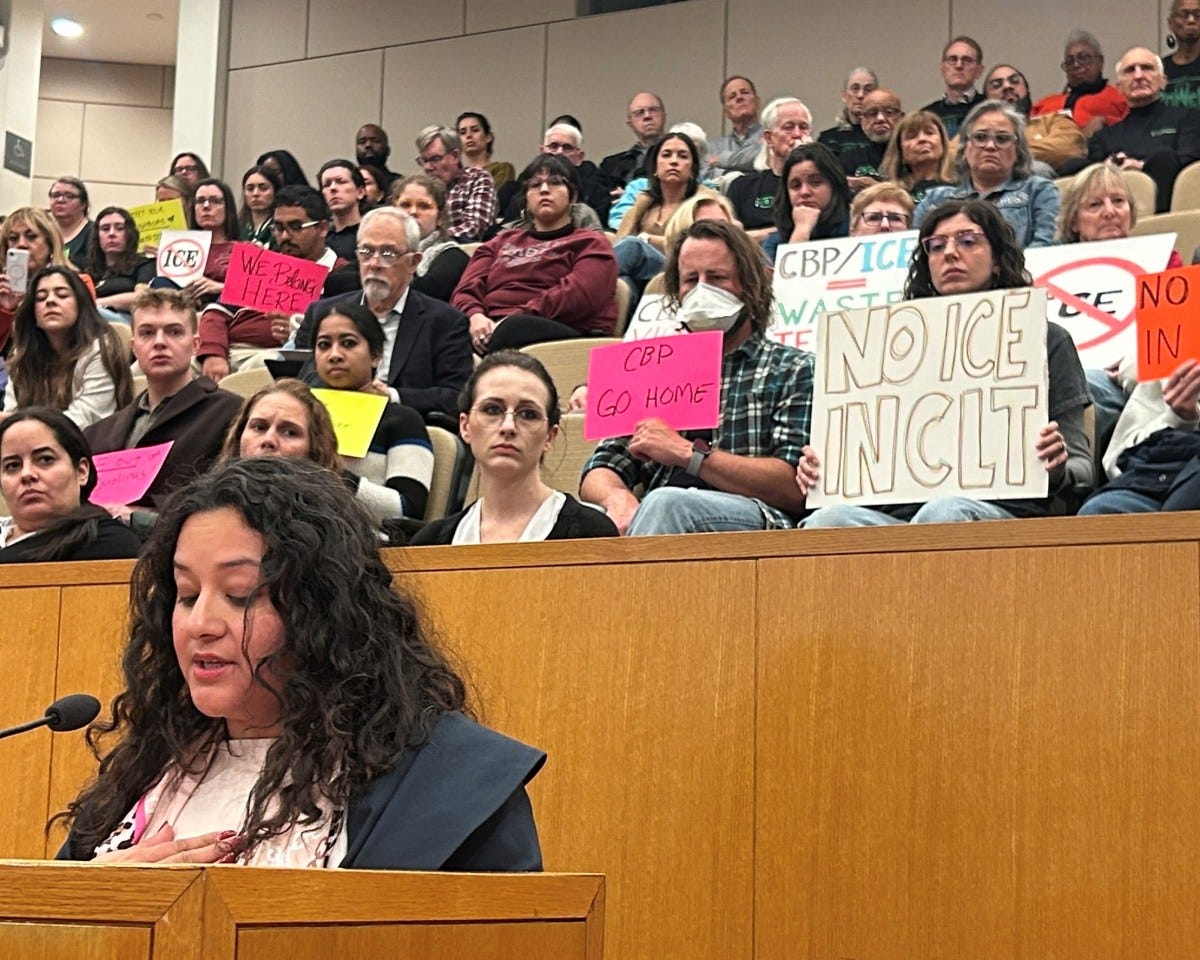A woman with black curly hair stands at a podium at an immigration protest with people holding signs behind her.