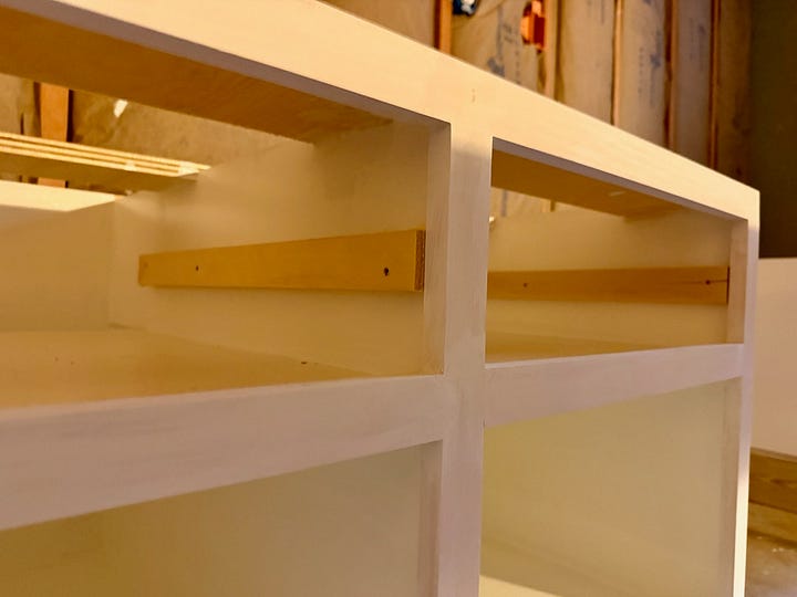 Two photos of basement built-in construction. The first shows cabinet bases with face frames installed beneath an unfinished ceiling. The second is a close-up view of freshly painted white face frames and interior shelving.