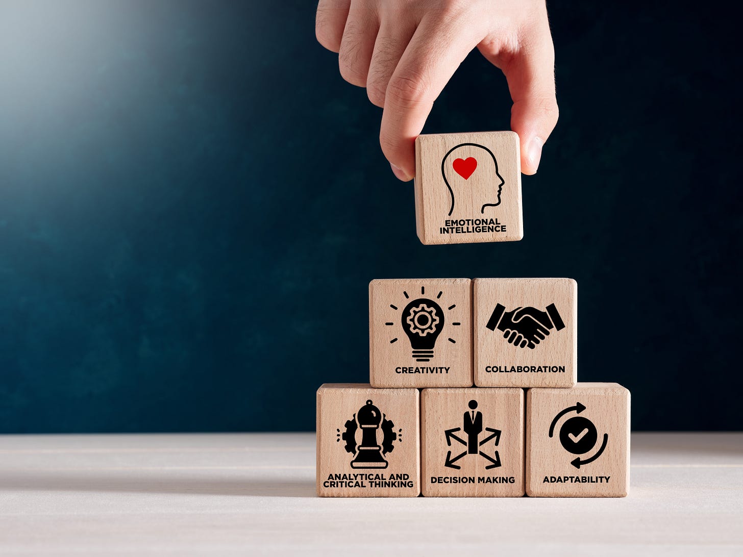 a hand placing a block that says emotional intelligence on top of a stack of other blocks that say creativity, collaboration, analytical thinking, decision making, and adaptability. There are graphic images on the blocks that correspond to those words.