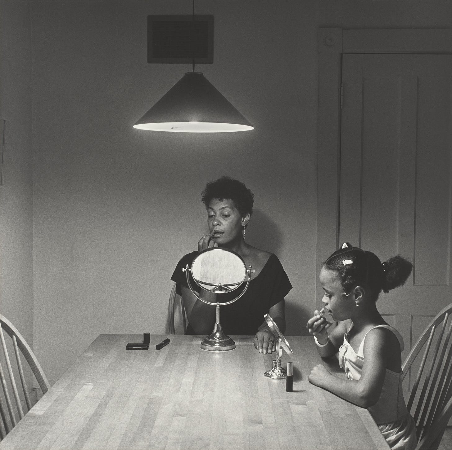 Black and white photo of a Black woman (Carrie Mae Weems) and Black girl sitting at a kitchen table, both with a small makeup mirror in front of them and applying makeup.