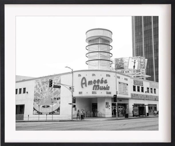 May include: A black and white photo of a building with a sign that reads 'Amoeba Music' in red letters. The building is located on a city street and has a large mural on the side. The photo is taken from a street level perspective.