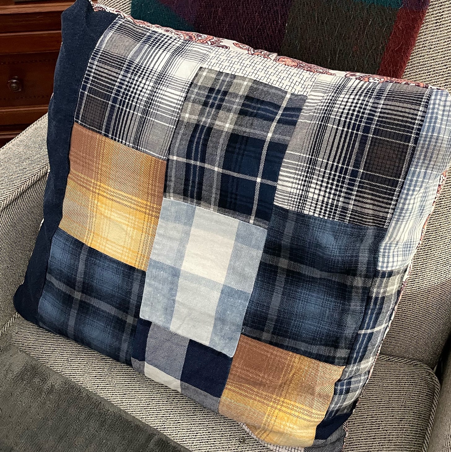 Square patchwork pillow with a plaid design in shades of blue, white, black, and mustard yellow, resting against a light gray upholstered chair. The pillow features a mix of different plaid patterns sewn together, creating a rustic, cozy aesthetic. A portion of a dark-colored woven blanket is visible in the background, adding to the warm and homey setting.