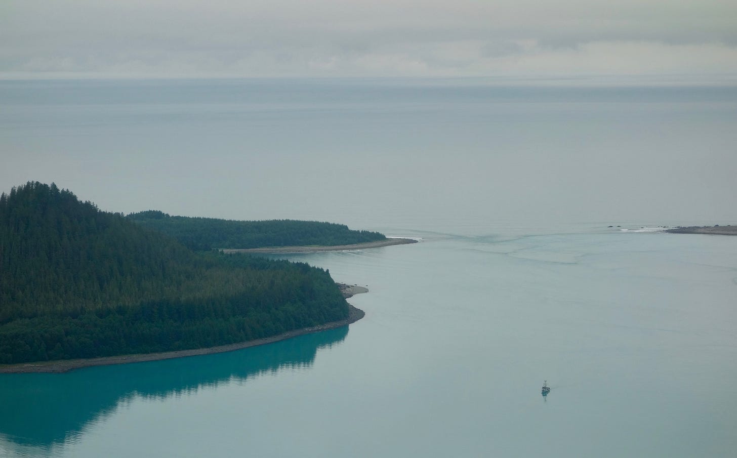 A boat sits in a calm bay, viewed from an aircraft. At the mouth of the bay, where it meets the ocean beyond, waves created by tidal currents are visible. A boat sits in a calm bay, viewed from an aircraft. At the mouth of the bay, where it meets the ocean beyond, waves created by tidal currents are visible.