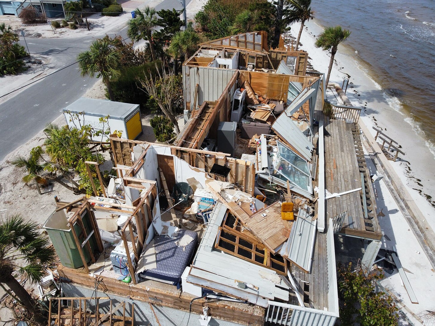daytime photo from above, showing a mostly-collapsed beachfront home with only some parts of its walls standing. The roof is gone entirely and furniture is strewn throughout the wreckage. daytime photo from above, showing a mostly-collapsed beachfront home with only some parts of its walls standing. The roof is gone entirely and furniture is strewn throughout the wreckage.