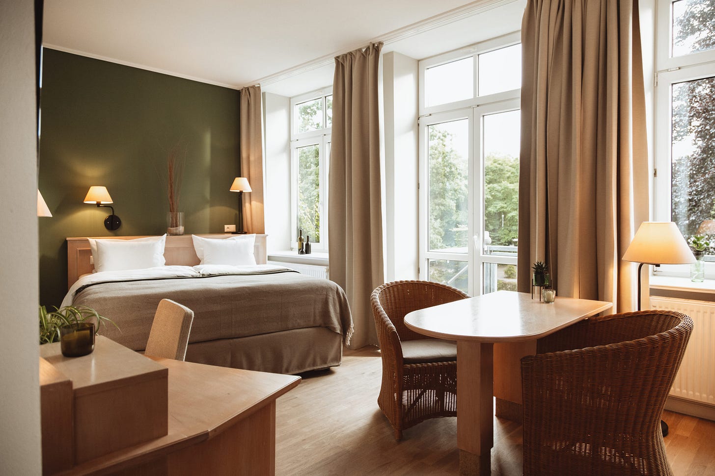 Hotel room with green walls, wooden furniture with natural bedding and curtains and large windows with balcony outside and green trees