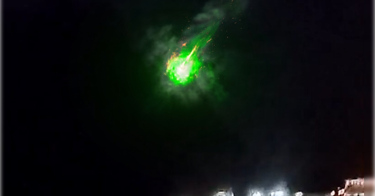 Bright green meteor streaking across a dark night sky, surrounded by glowing clouds and sparks, with illuminated vehicles below.