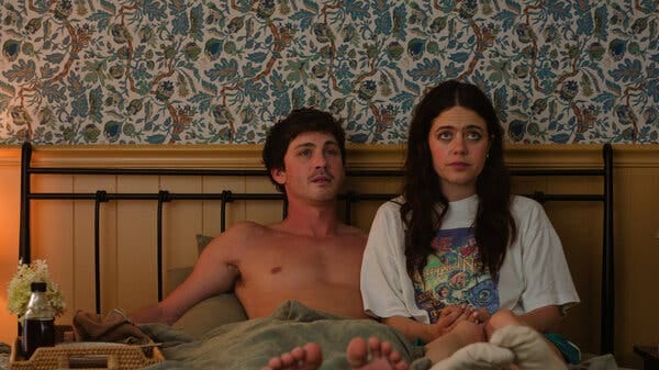 A shirtless man and a woman in a graphic T-shirt sit upright in bed against a floral wallpapered wall, both looking ahead with serious expressions.