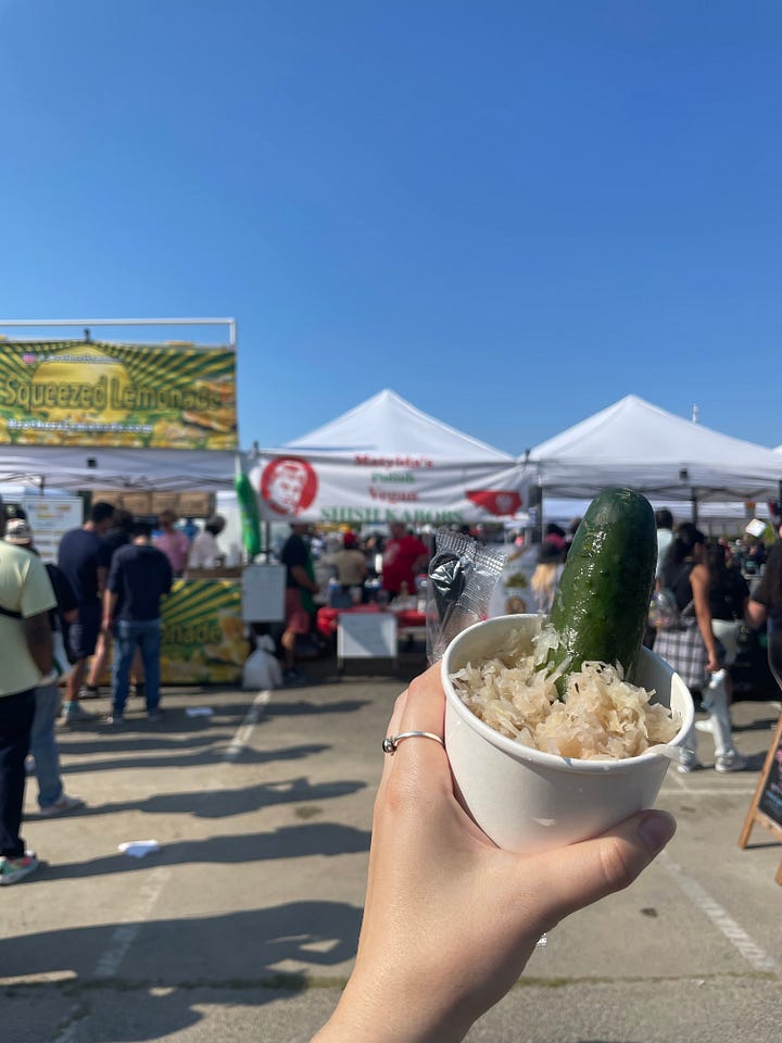 man holds cup of purple ice cream and coconut cream with red beans at the bottom, a pickle and some sauerkraut in front of a food tent