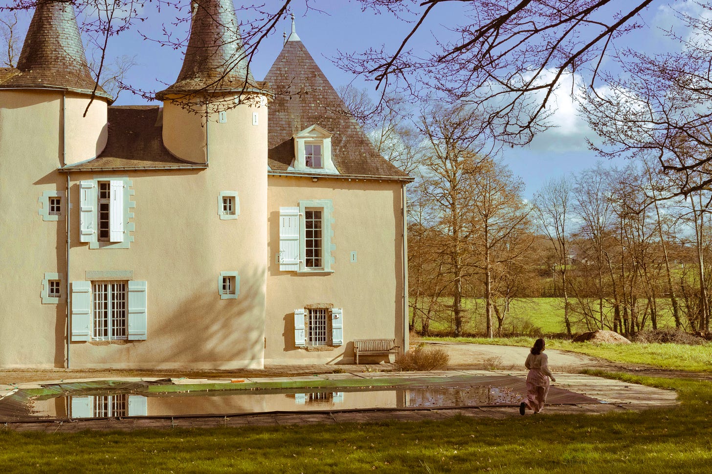A woman in a long dress walking across the lawn past a covered swimming pool, with the château and its turrets rising behind her in afternoon light. A woman in a long dress walking across the lawn past a covered swimming pool, with the château and its turrets rising behind her in afternoon light.