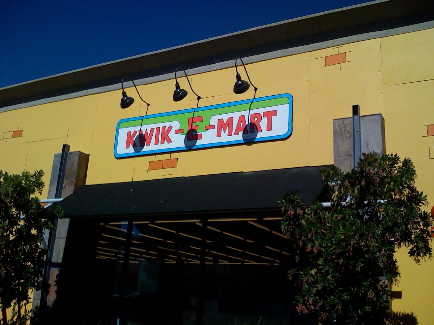 The entrance to a 7-Eleven store on Pear Avenue in Mountain View, California that is dressed as a Kwik-E-Mart. The sign above the entrance reads Kwik-E-Mart. The building is covered in yellow foam core to complete the transformation.