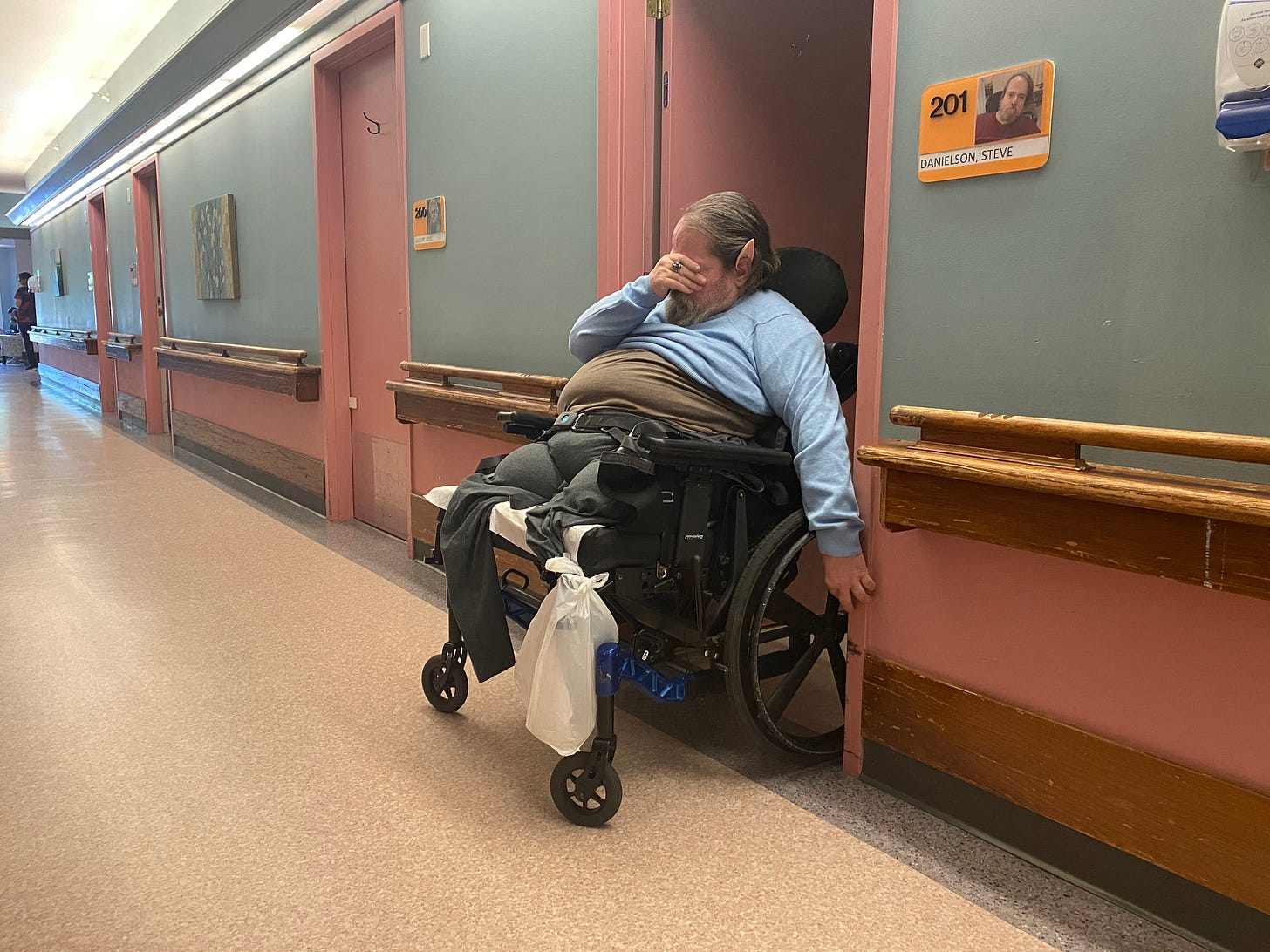 A man in a wheelchair is positioned in the doorway of a room in a care home. A plaque on the wall near the door displays his image, room number, and name. He is wearing a blue sweater and Vulcan “Spock ears,” with one hand covering his eyes.