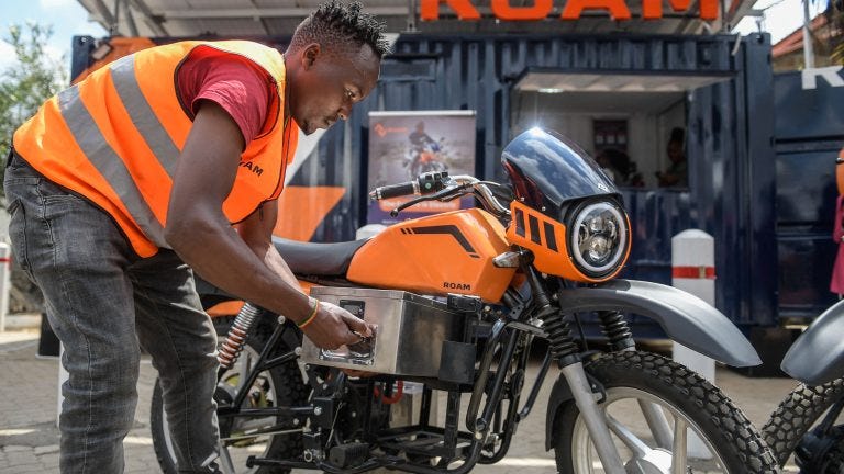 A photograph showing a man removing a battery from an orange, electric motorbike. A photograph showing a man removing a battery from an orange, electric motorbike.