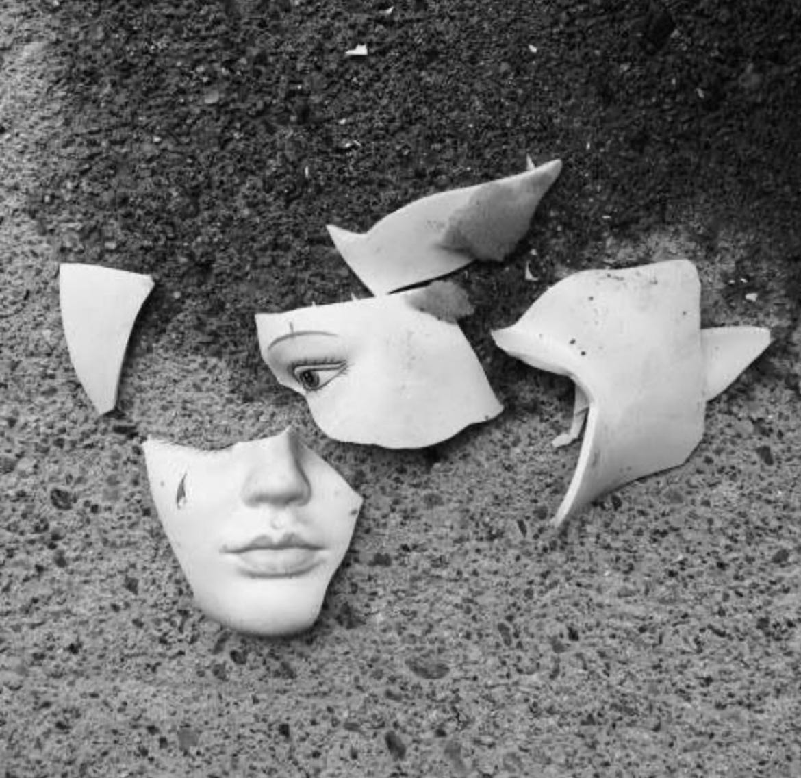 Photo of a shattered white porcelain or ceramic piece against the grey backdrop of the ground. Some pieces of the shattered object appear to feature a face; one piece has an eye and an eyebrow, another has a nose and a mouth.