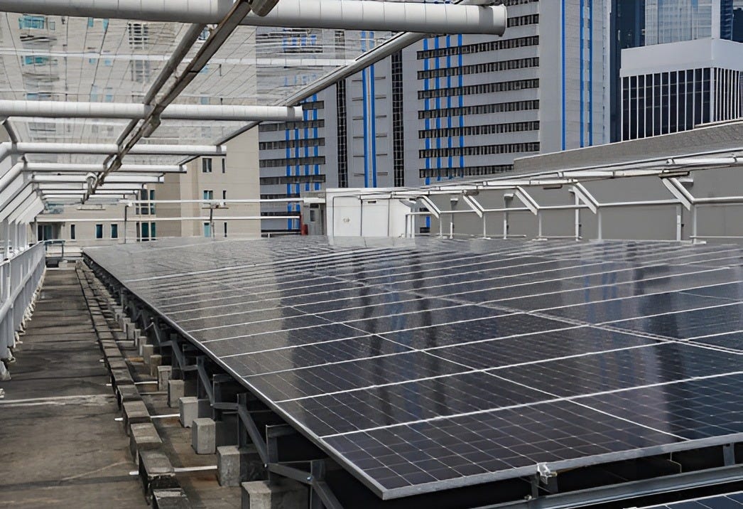 A high-angle, wide shot showing a large installation of sleek, dark-blue solar panels covering the rooftop of a modern building in an urban setting. The panels are mounted on a sturdy metal frame, with white industrial piping and railings running alongside the installation. In the background, tall, contemporary skyscrapers with glass and blue-tinted facades rise under a clear sky, illustrating the integration of renewable energy infrastructure within a dense city center like Jakarta.