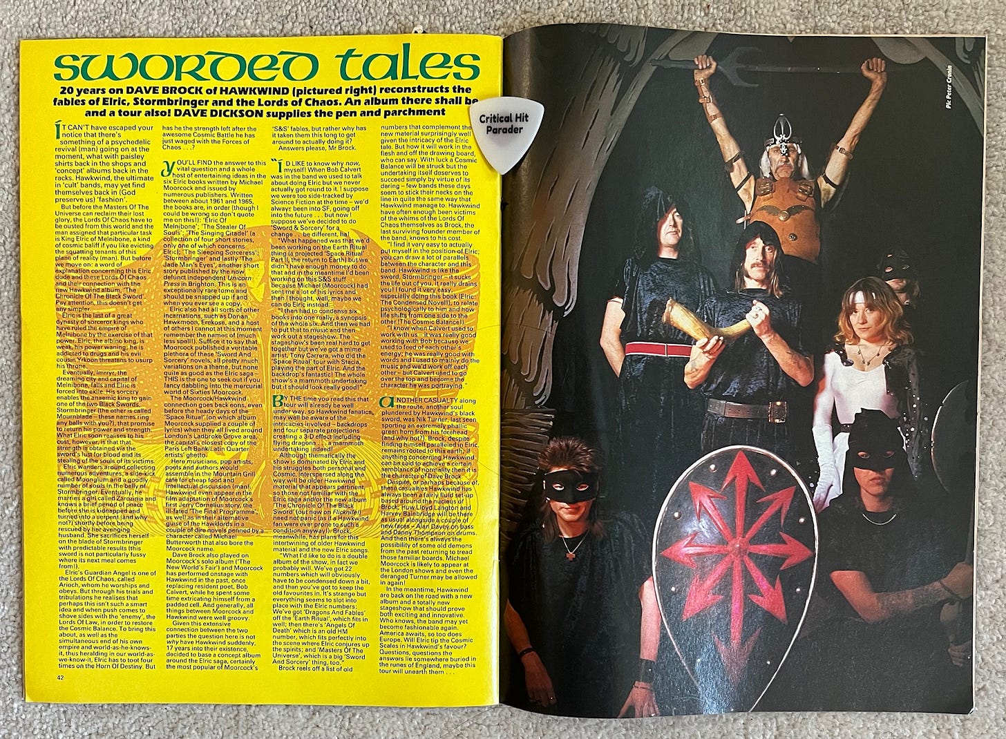 Two facing pages from Kerrang magazine. The left page has a Hawkwind article titled "Sworded Tales." It is on a yellow background with a red background image that makes the article tough to read for this colorblind person with old eyes. The right page is a photo of Hawkwind dressed in various hoods, masks and helmets with two well-mustachioed members holding a horn and sword respectively. In front of the band is a large black shield with the 8-arrowed symbol of Chaos in red.