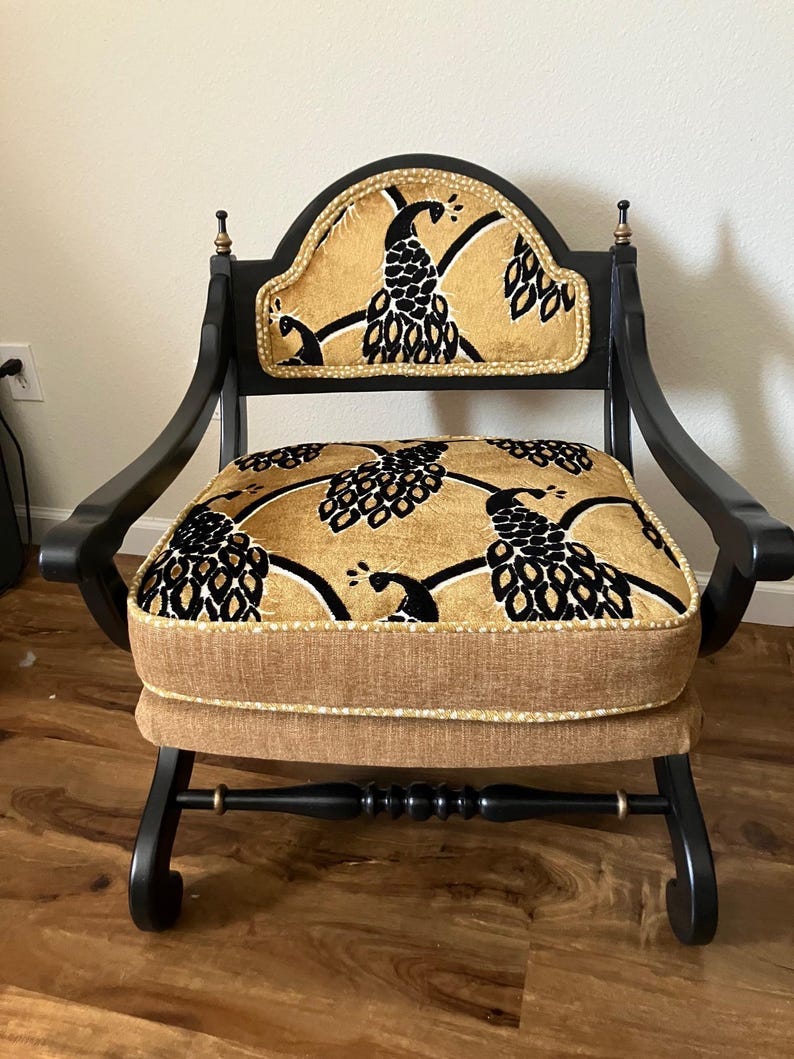 May include: An antique black armchair with a decorative peacock pattern on the seat and backrest. The chair features a curved back, armrests, and a cushioned seat. The fabric is gold with black peacock designs, and the chair has gold accents.