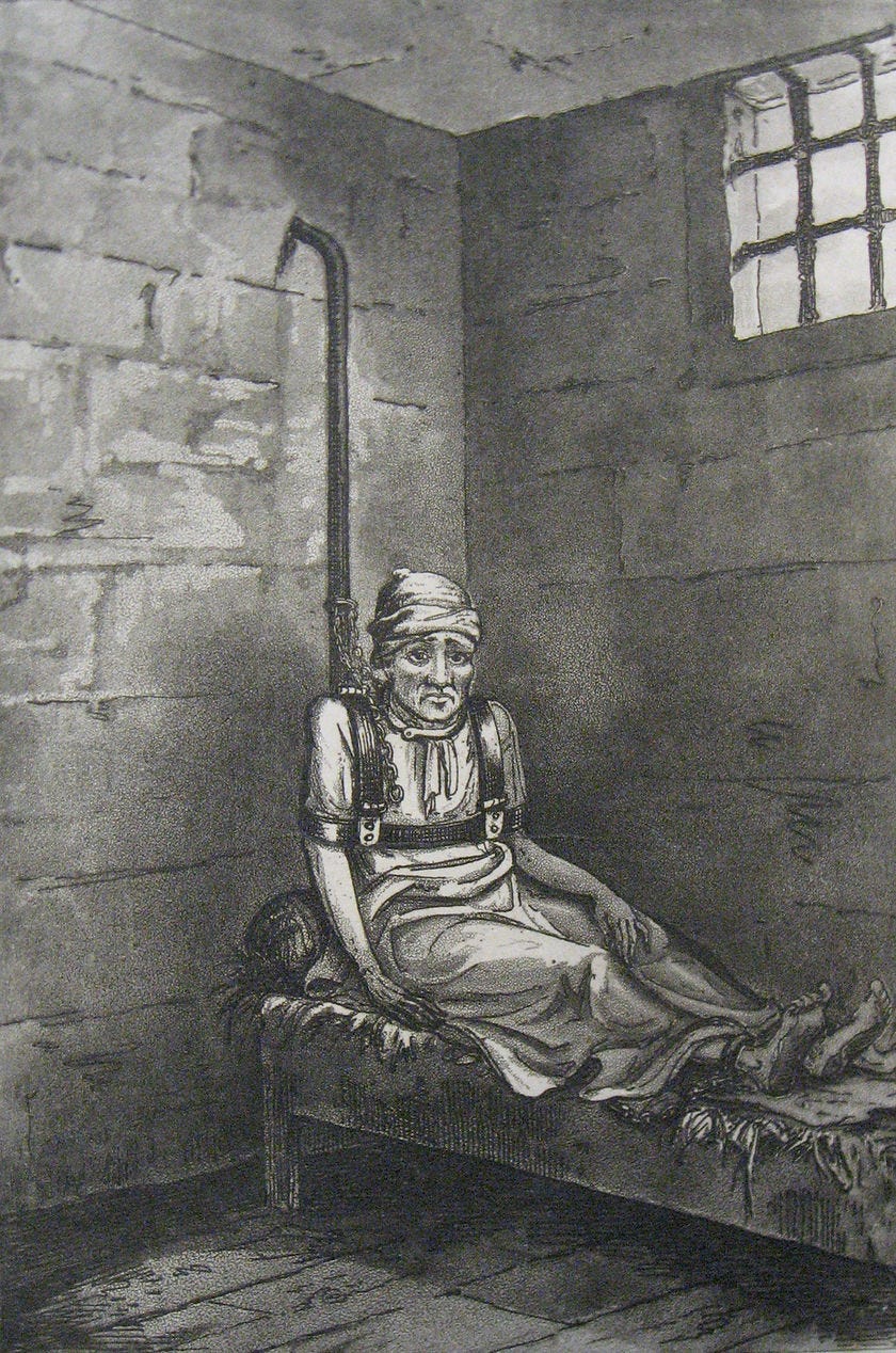 William Norris is shown in a dark cell frowning, strapped to a pole in bed