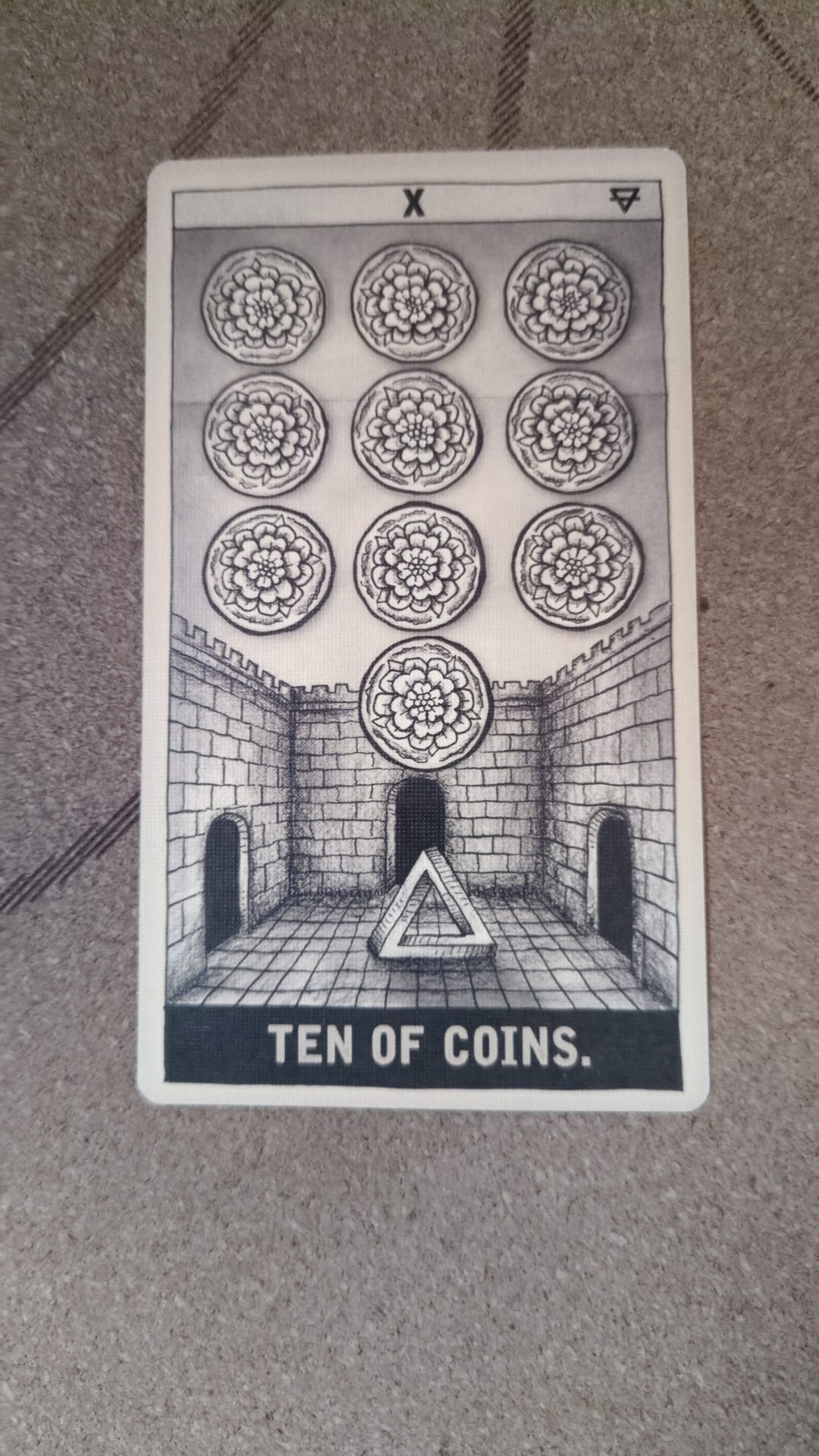 A black and white tarot card showing the Ten of Coins. Ten floral coins are arranged in a grid above a stone courtyard enclosed by walls, with three dark arched doorways. A single coin is centred above the middle doorway, and a triangular symbol rests on the tiled floor below. The card has a stark, architectural feel and is labelled “TEN OF COINS.”