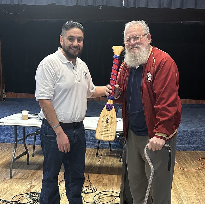 Two men shake hands in a wood‑paneled hall near a small stage. The man on the left wears a white polo and jeans; the older man on the right, with a white beard and glasses, wears a red jacket and gray slacks and leans on a decorative cane wrapped with red‑and‑blue cord. A small paddle attached to the cane bears an insignia with a dome resembling the U.S. Capitol and the words “Commanders Coaliiton.” A black Behringer PA speaker sits between them, with audio cables running to a folding table holding equipment. A blue flag with gold fringe and framed plaques hang on the wall in the background.