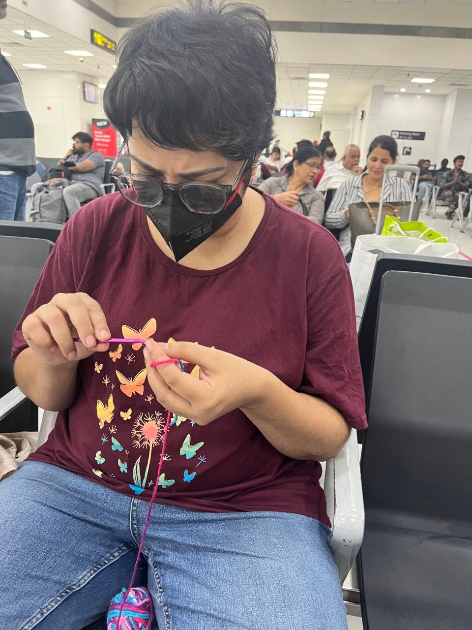 I am seated at the airport dressed in a red top with a mask and glasses. I am focussing on a stitch with a ball of yarn in my lap and a hook in my hand. 