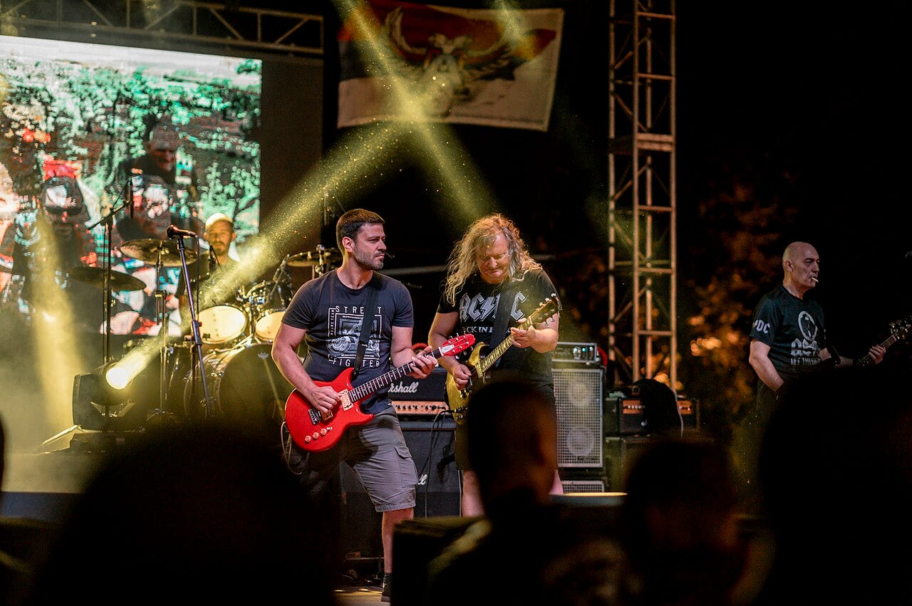 Photo: Shixart1985 / Wikimedia Commons (CC BY 2.0) Musicians perform energetically with electric guitars on stage at a live rock concert, illuminated by colorful lights while the audience enjoys the performance at night.
