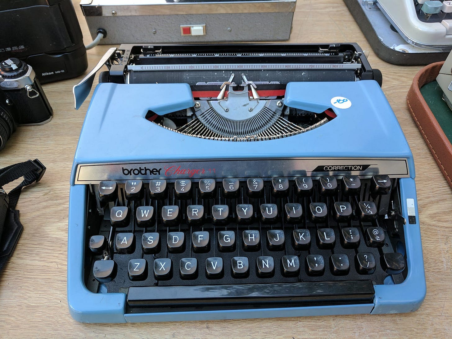 A sky-blue Brother Charger typewriter on a wooden table, flanked by vintage cameras. A sky-blue Brother Charger typewriter on a wooden table, flanked by vintage cameras.