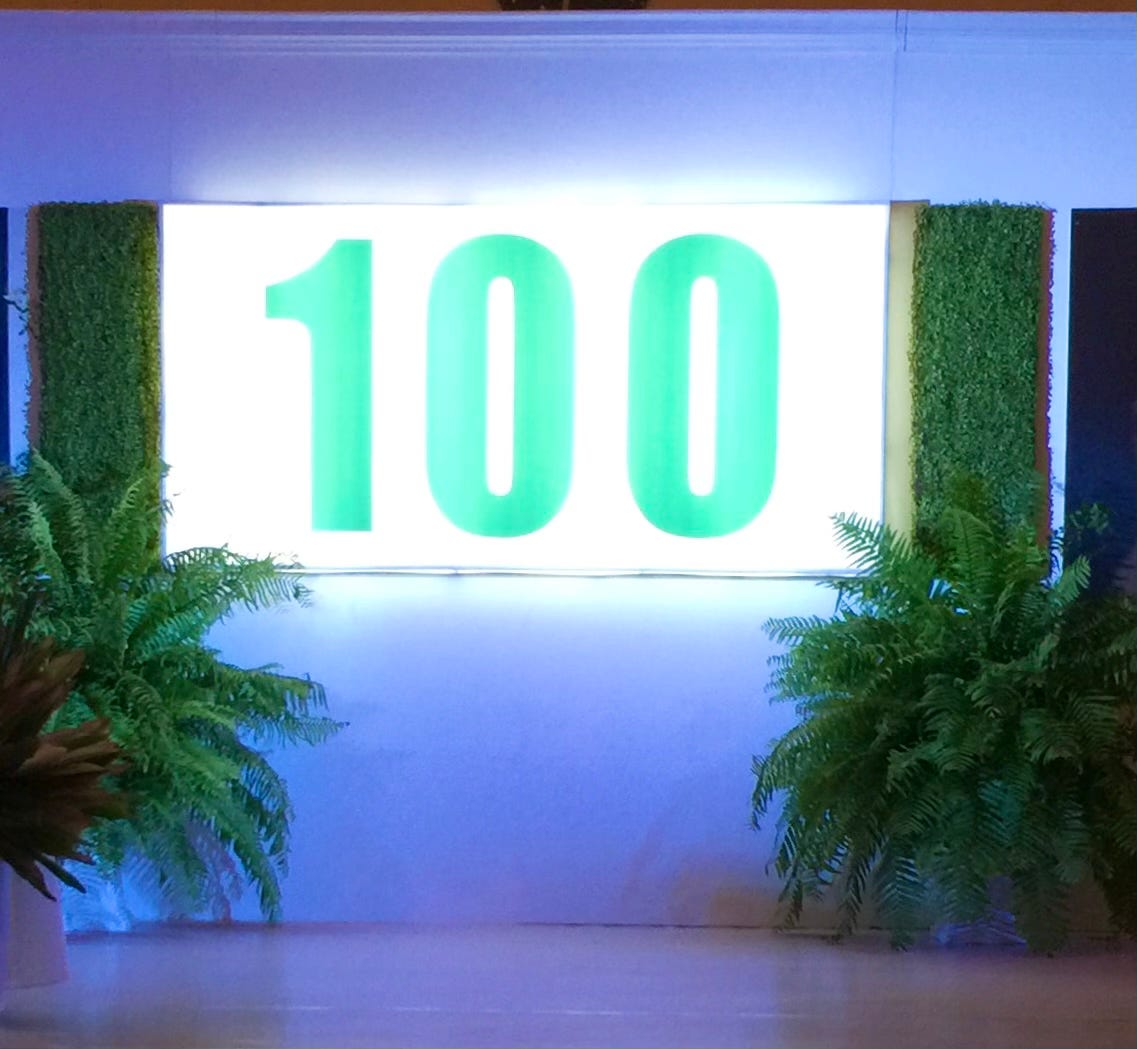 The number 100 giant in screen over a neon board. Pots of ferns on the floor nearby.