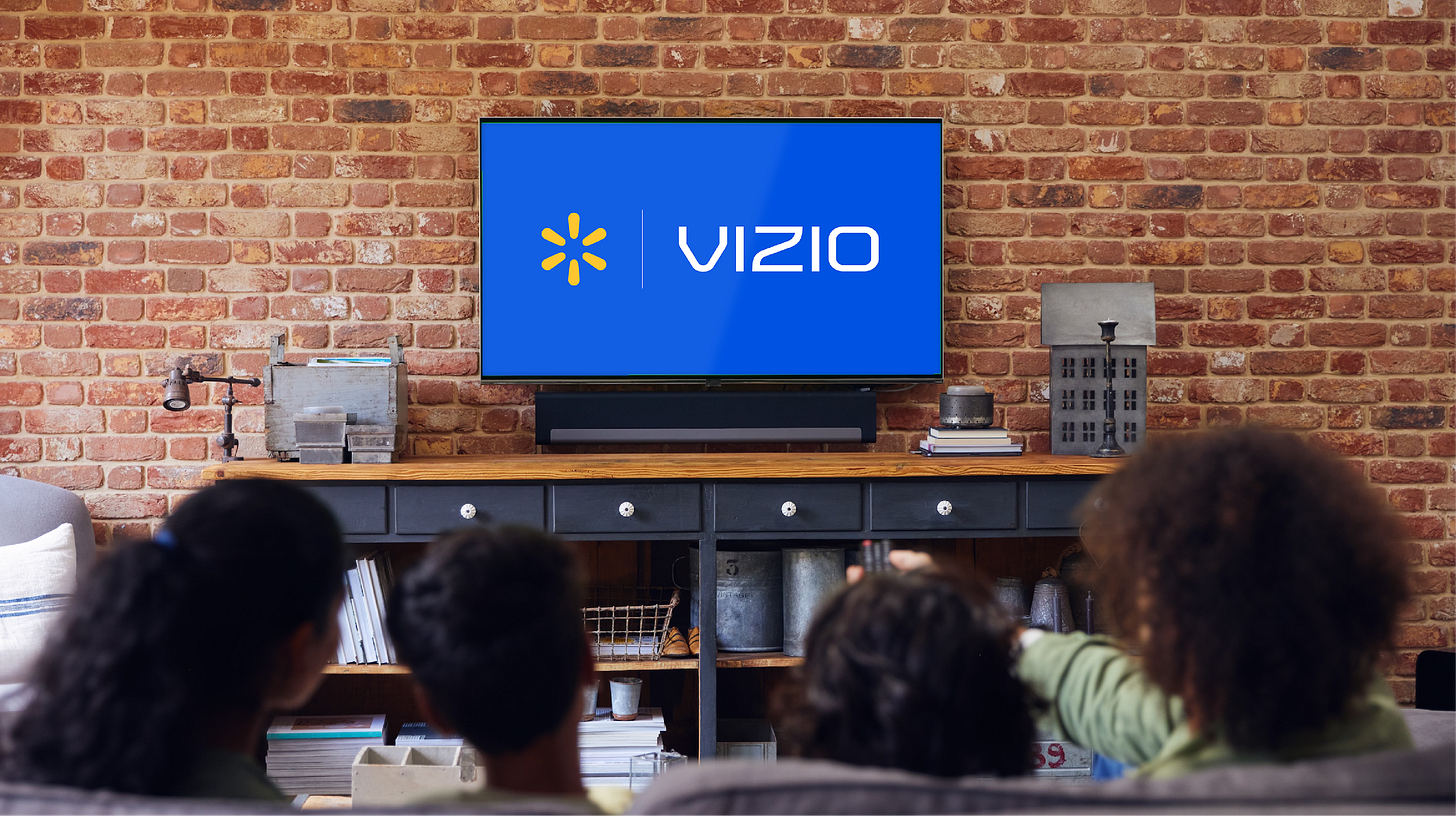 A group of four people sits together on a couch, watching a Vizio television in a home living room. The TV screen displays the unmistakable Vizio logo on a blue background. The setting features a brick wall, wooden console, and modern decor, creating a warm and inviting atmosphere. The group includes adults and children, viewed from behind, suggesting a family movie or TV night. A group of four people sits together on a couch, watching a Vizio television in a home living room. The TV screen displays the unmistakable Vizio logo on a blue background. The setting features a brick wall, wooden console, and modern decor, creating a warm and inviting atmosphere. The group includes adults and children, viewed from behind, suggesting a family movie or TV night.