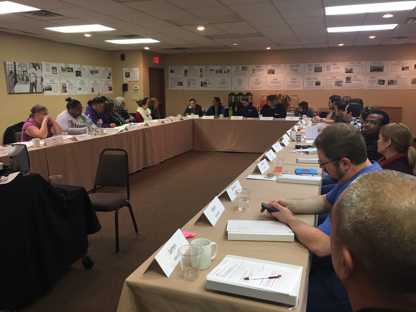 people are sitting at a large U shaped table formation with name plates in front of them and posters lining the wall