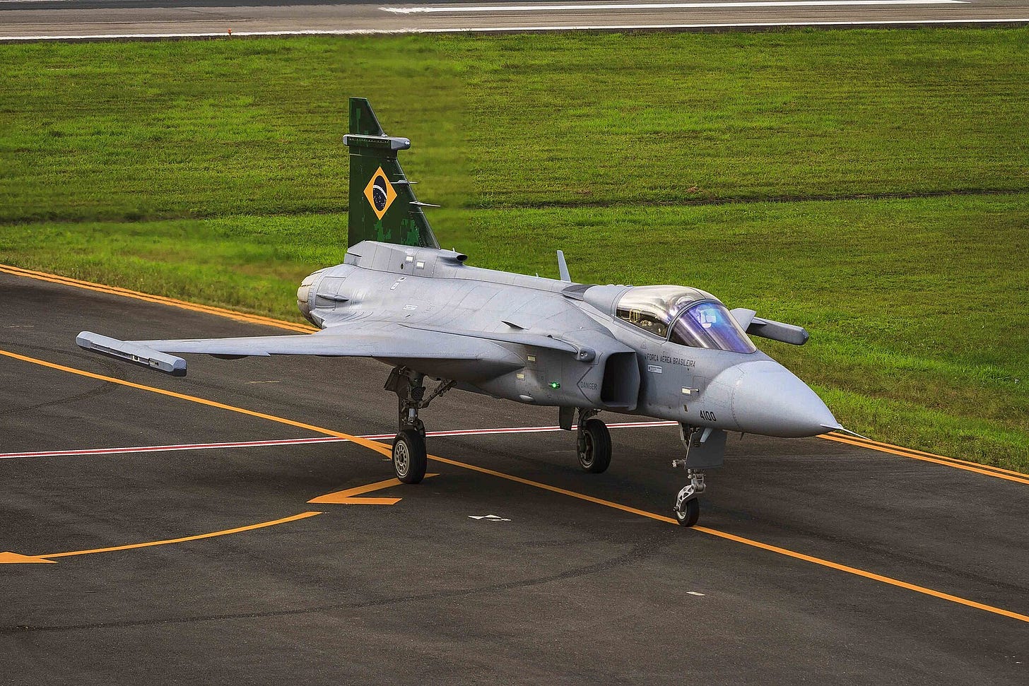 A grey Gripen fighter with a green and yellow tail emblem taxiing on a runway next to green grass.