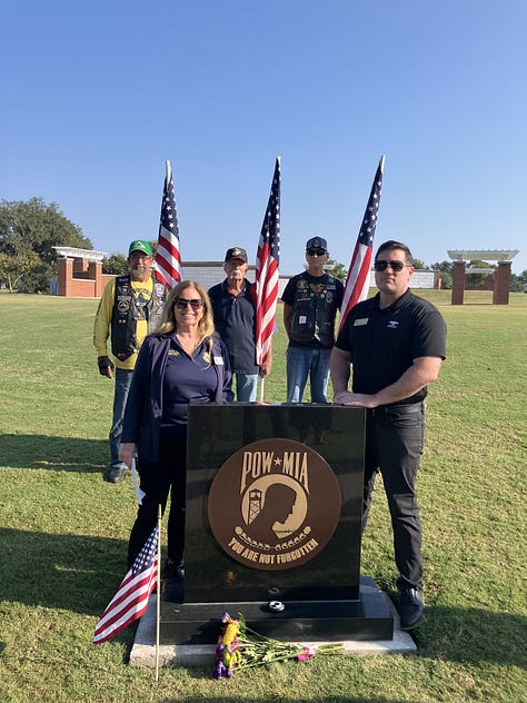 A group of veterans and community members pose outdoors at a POW*MIA memorial. In the foreground is a black granite monument engraved with the POW*MIA emblem and the words “YOU ARE NOT FORGOTTEN,” with a small American flag at its base. Several U.S. flags stand behind the group. Some people wear military dress uniforms and VFW-style caps; one cap reads “Tallahassee,” and another hat bears the U.S. Air Force insignia. On the right, an elderly veteran in a wheelchair salutes while a man in a suit speaks with him. The scene is on a sunny day in a grassy field with cars and flagpoles in the background.