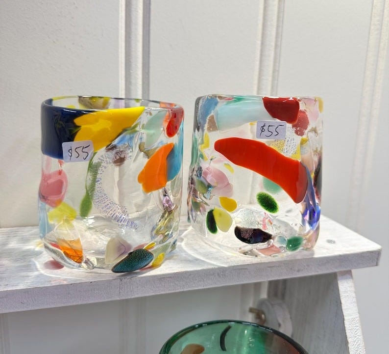 May include: Two clear glass cups with colorful glass pieces embedded in the glass. The cups are on a white shelf. The cups are priced at $55 each.