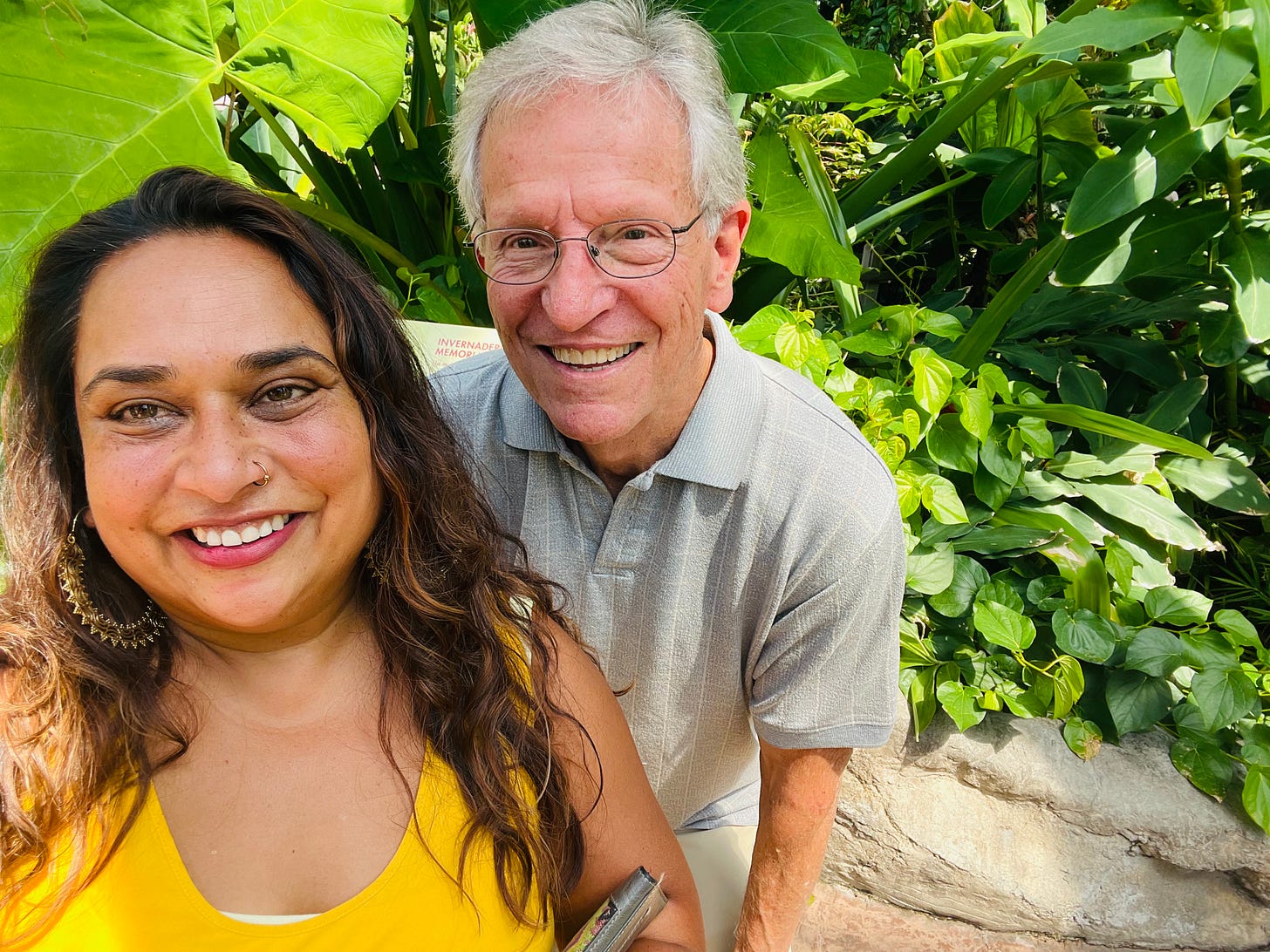 Selfie of Vini and Paul in a garden. Vini is in the foreground smiling, with a yellow shirt on and gold hoop earings. Paul is behind her with a grey collared shirt, white skin, grey hair, glasses and a big smile.