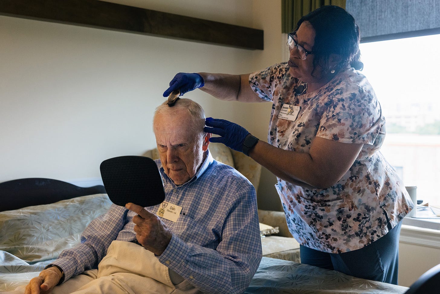 A senior man holds a mirror to watch as a nurse brushes his short, white hair.
