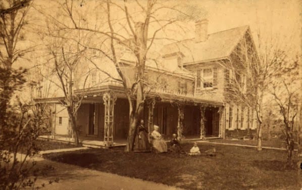 Sepia image of two-story house with large porch; four figures are on the lawn, next to a tree. Sepia image of two-story house with large porch; four figures are on the lawn, next to a tree.