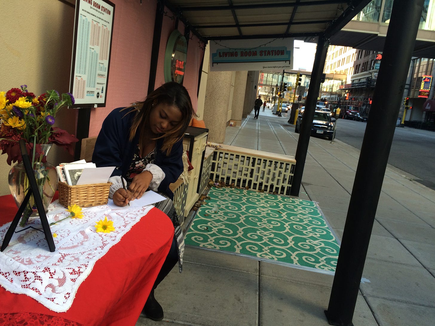 A woman sits at a cheerful red table with a vase of flowers on it. Behind her is another seating area with a teal rug and benches with pillows on them.