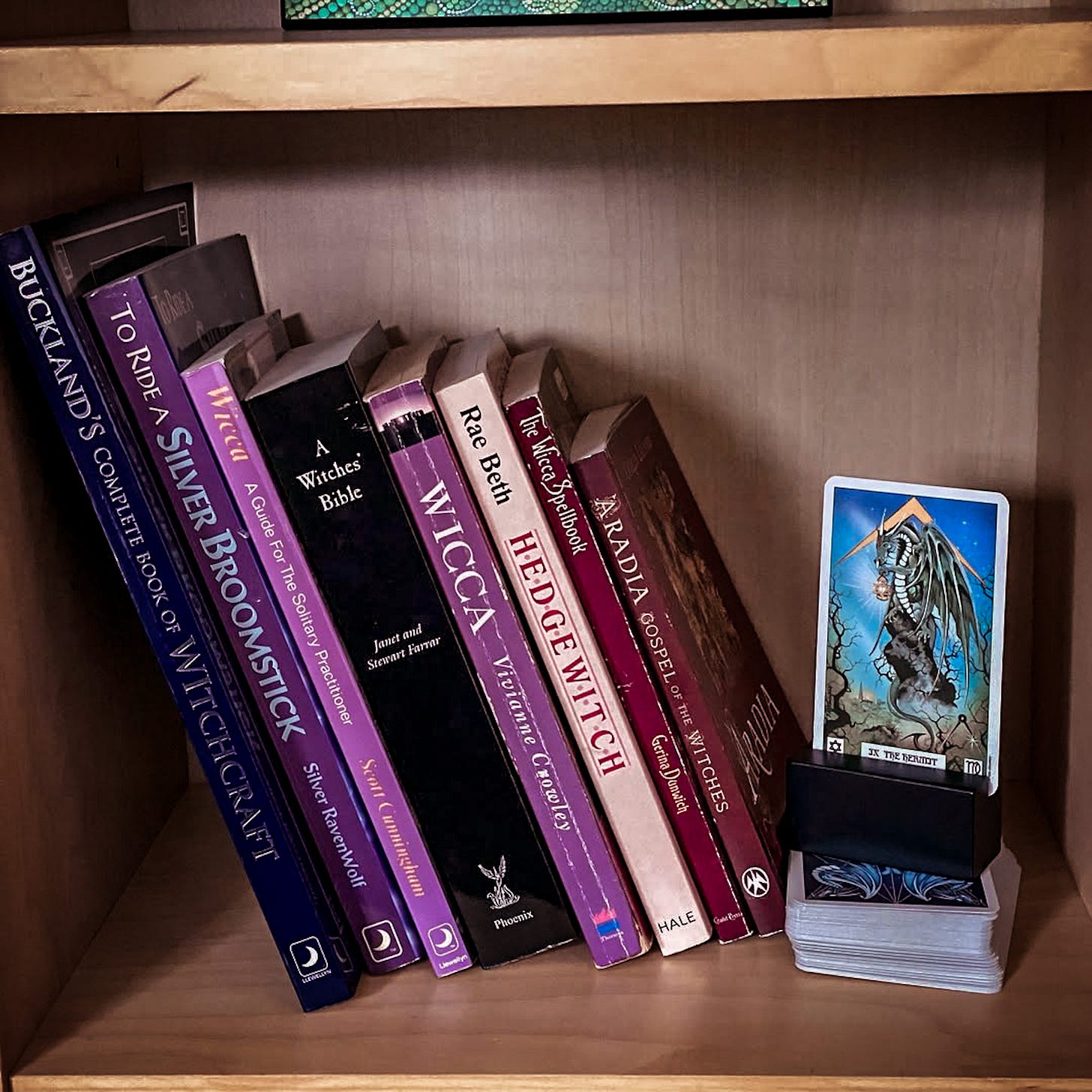 Several books are shown on a bookshelf: Buckland's Complete Book of Witchcraft; To Ride a Silver Broomstick; Wicca: A Guide for the Solitary Practitioner; A Witches' Bible; Wicca: The Old Religion in the New Millennium; Hedge Witch; The Wicca Spellbook; and Aradia: Gospel of the Witches. The Hermit Card from Peter Pracownik's Dragon Tarot is also shown upright, in a card holder on top of the rest of the deck.