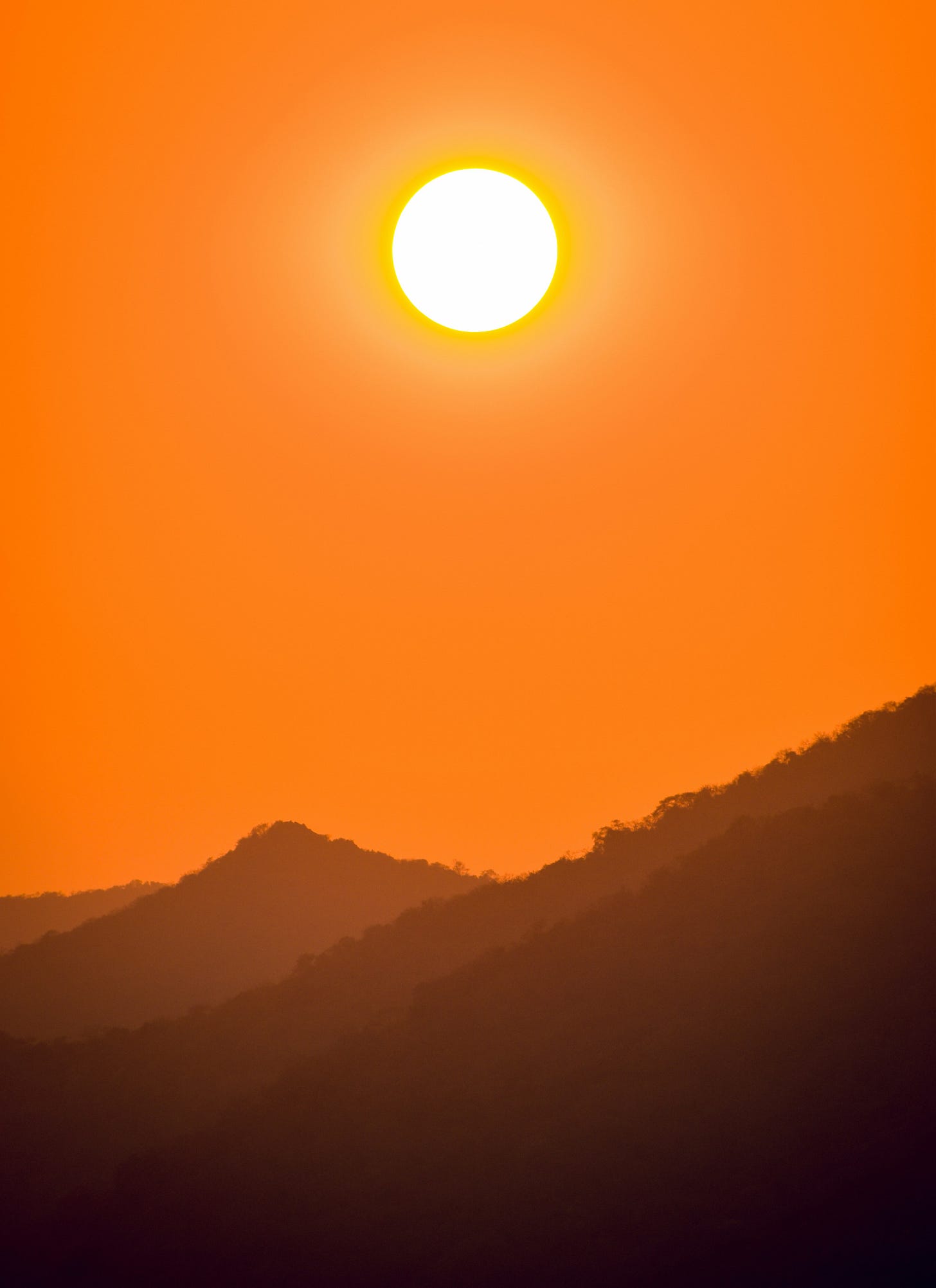 The sun beating down on mountains. The sky is orange and there is haze in the foreground, blurring the mountains in the back. The sun beating down on mountains. The sky is orange and there is haze in the foreground, blurring the mountains in the back.