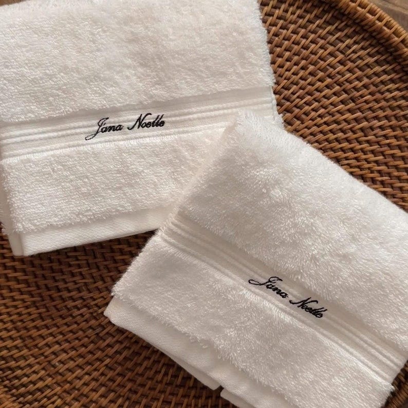 May include: Two white bath towels with embroidered text. The towels are folded and have a textured appearance. The embroidered text on each towel reads 'Jana Noelle' in a cursive font. The towels are placed on a woven, brown surface.
