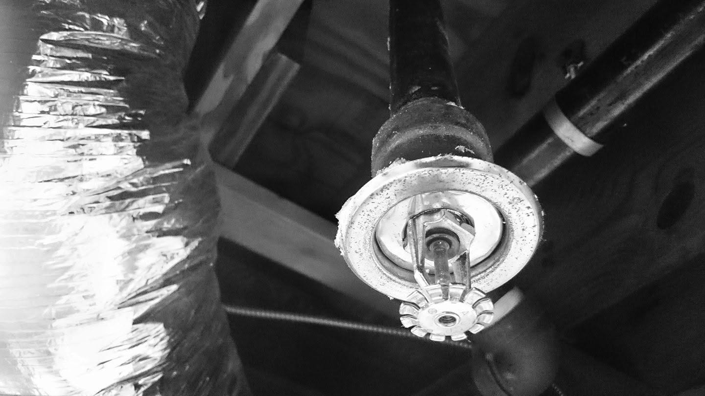 Black-and-white photograph looking upward at a fire sprinkler head surrounded by exposed pipes and reflective ductwork.