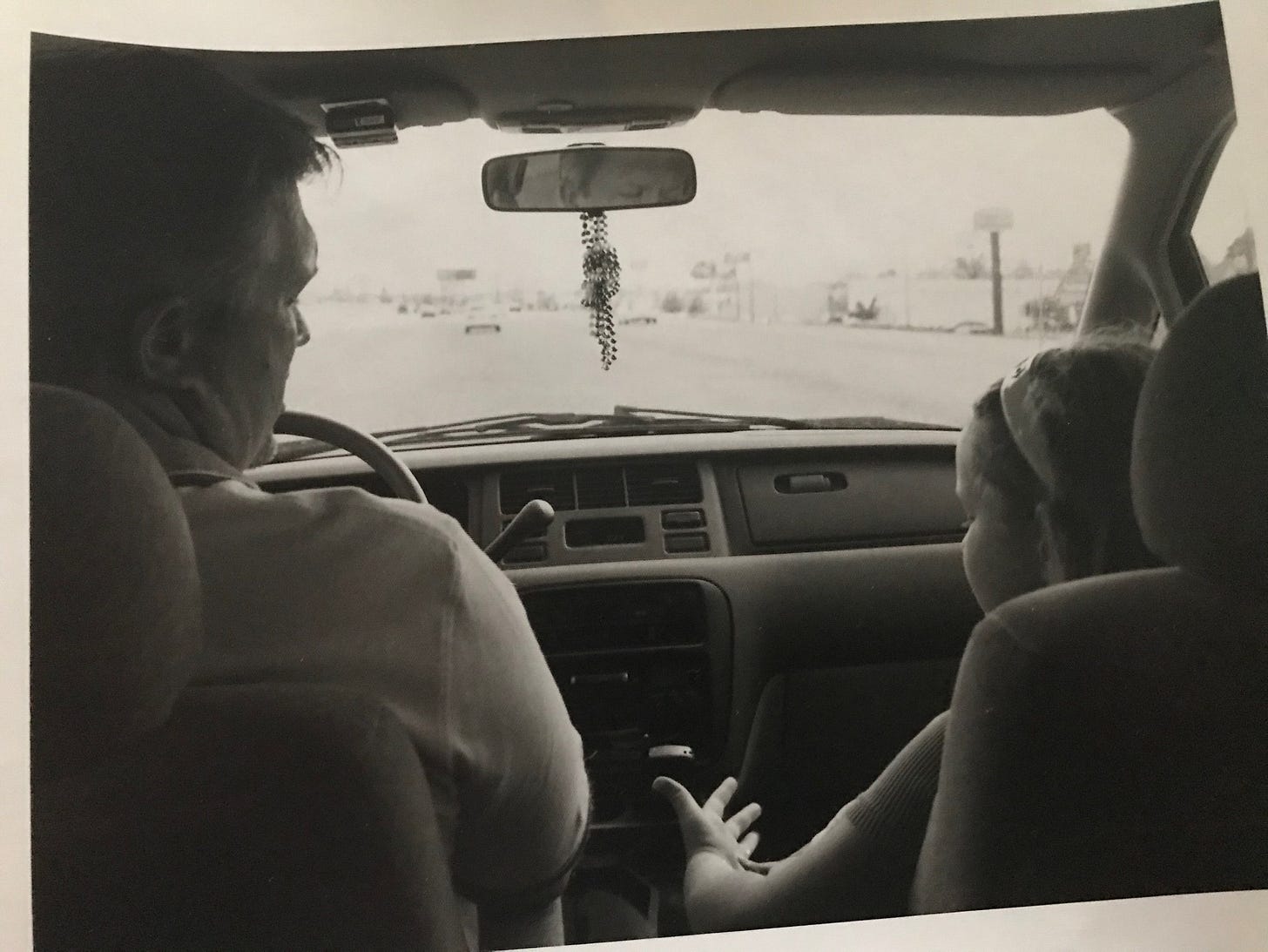 Krys talking about something in the front seat of the car while their dad looks on and listens. Picture is in black and white and features a late 90s car interior.
