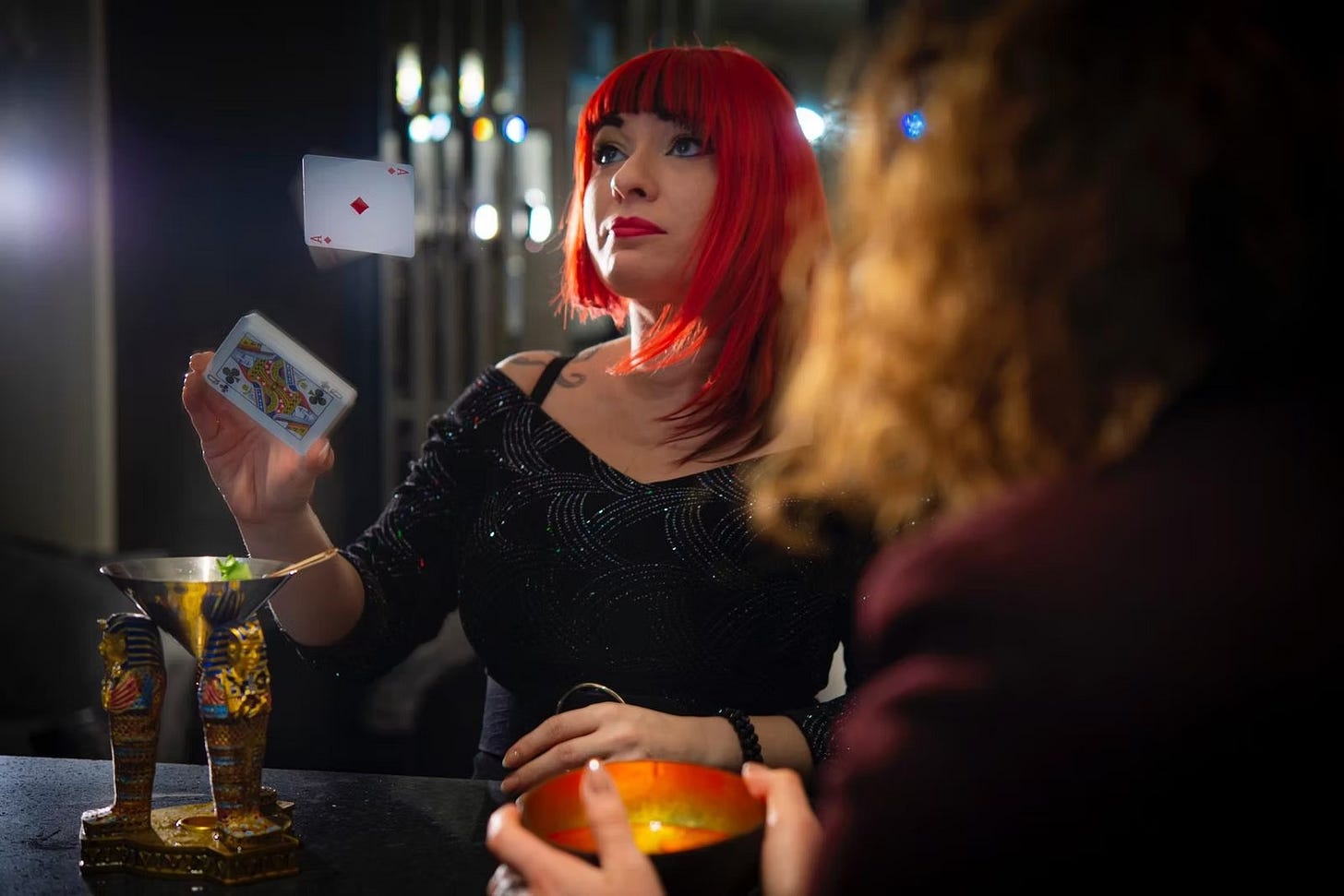 A woman with bright red hair wearing a sparkly black top performs a magic trick at a dimly lit bar, looking up as an Ace of Diamonds playing card flies through the air above the deck in her hand. A spectator in the foreground watches the performance.