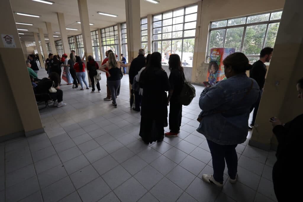 People stand in a long line inside a tiled, covered corridor, waiting to enter a building People stand in a long line inside a tiled, covered corridor, waiting to enter a building