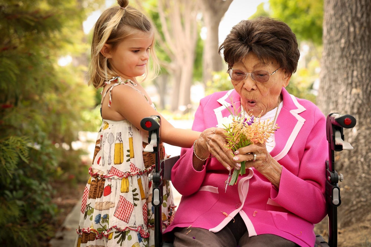 A young girl in a colorful patterned dress stands beside an elderly woman in a bright pink blazer who is seated in a wheelchair. The girl gently rests her hand on the wheelchair as the older woman holds and smells a small bouquet of wildflowers. They are outdoors in a park or garden setting with dappled sunlight filtering through trees in the background.