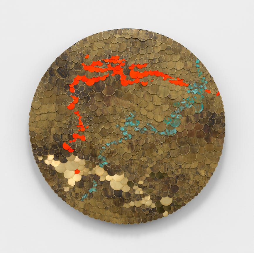 A circular wall-based artwork made of small metallic disc-like elements, with abstract red and turquoise markings across a gold-toned surface.